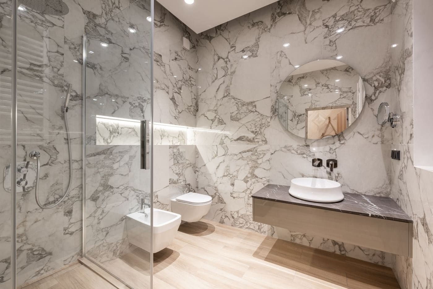 A bathroom lined in white marble-look tiles with a round mirror and a floating vanity