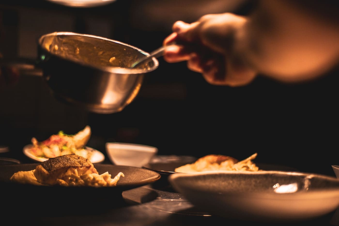 A cook spooning sauce over plates on a dark kitchen pass
