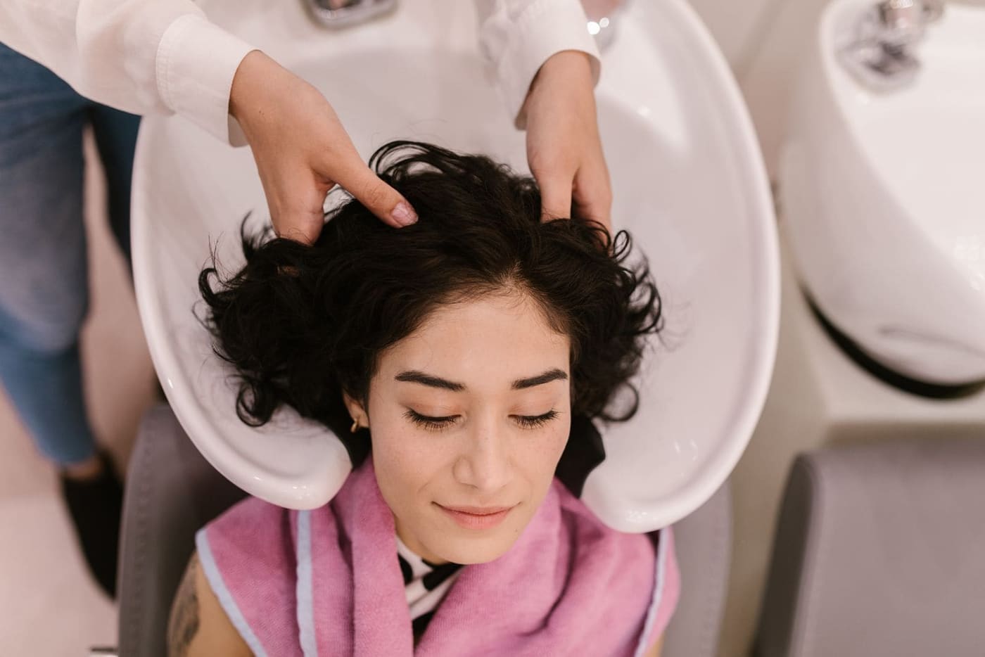 A woman with dark curly hair relaxing at a wash basin while a stylist massages her scalp