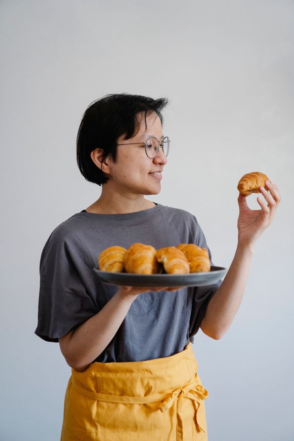 A smiling baker in glasses holding a tray of fresh rolls and a croissant