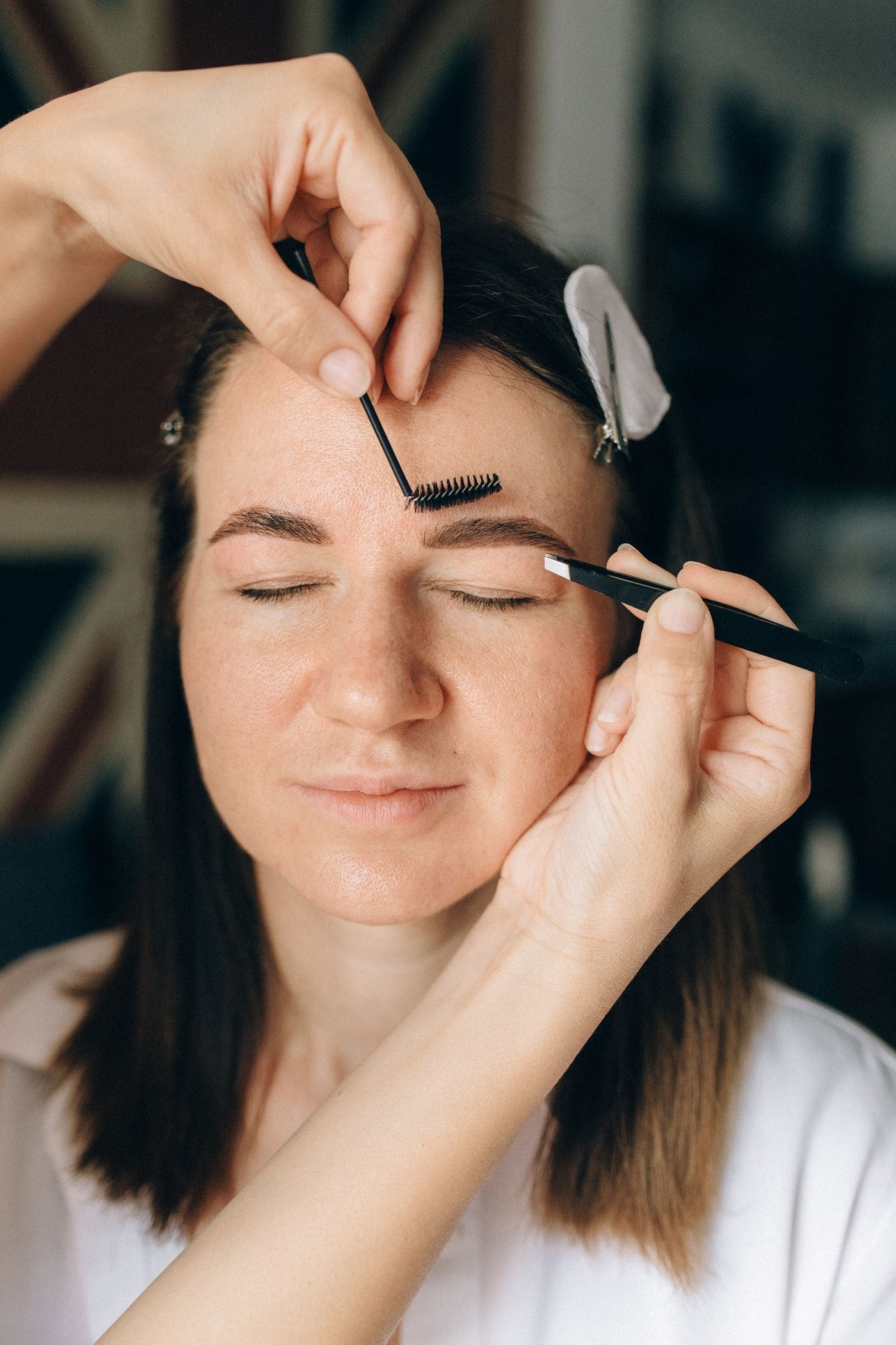 A brow artist shaping a client's eyebrow with a spoolie and tweezers