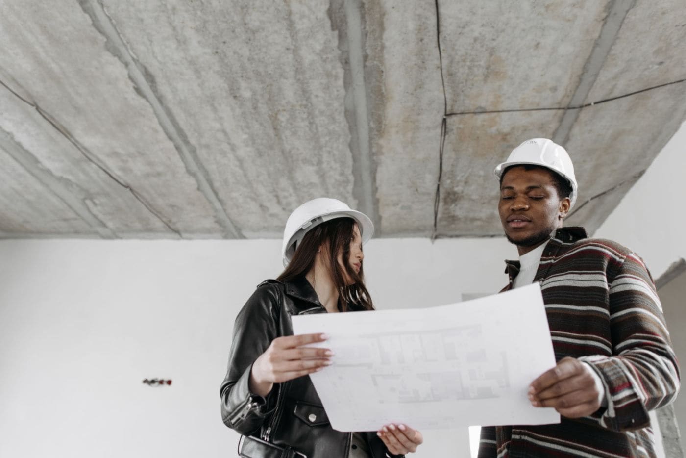 Two builders in hard hats reading a plan inside a house being renovated