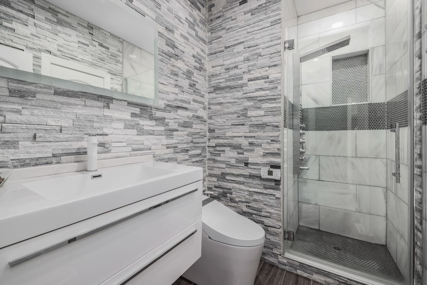 A bathroom with stacked stone tiles, a floating white vanity and a glass shower