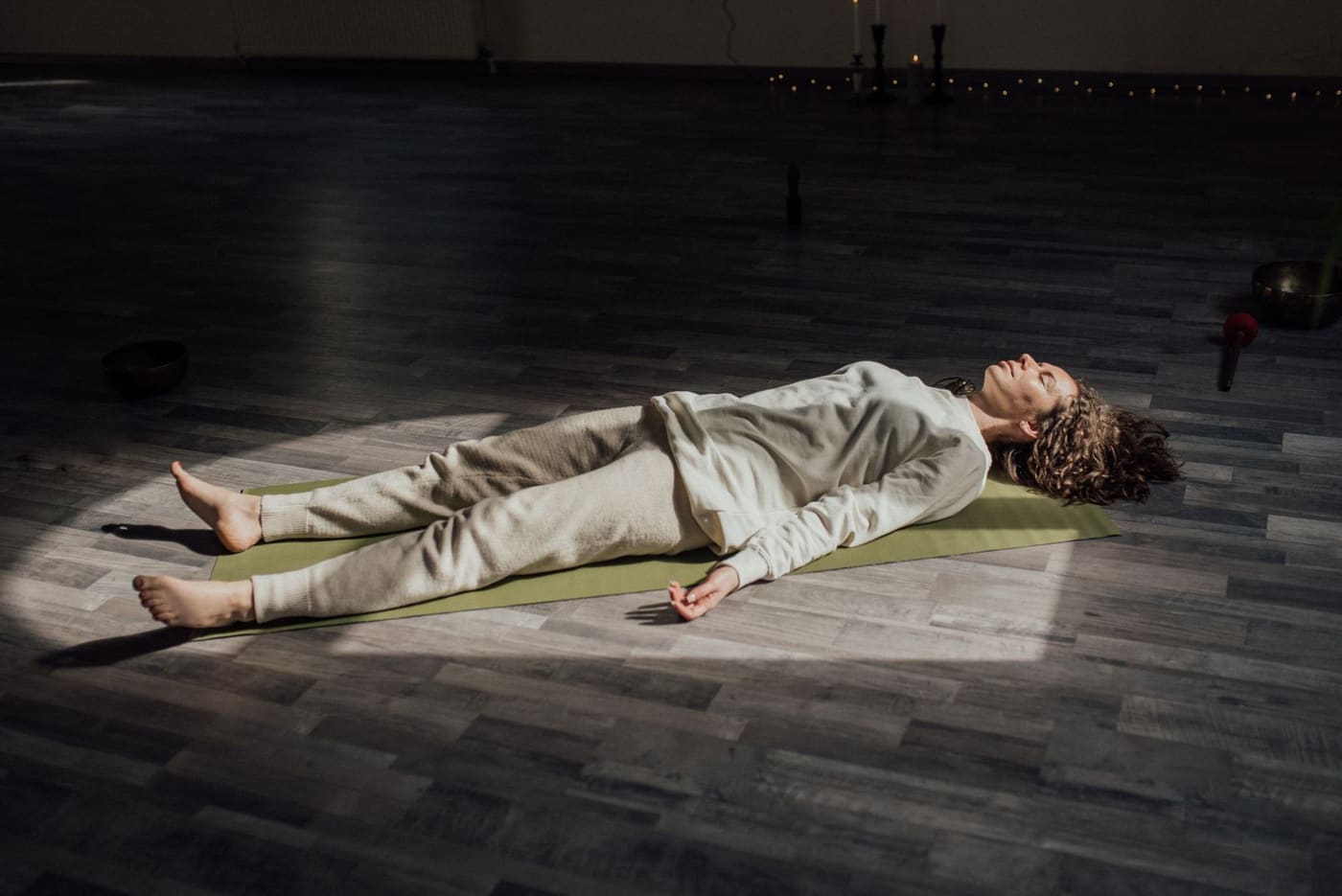 A woman lying still on a mat in a dark studio, lit by a patch of window light