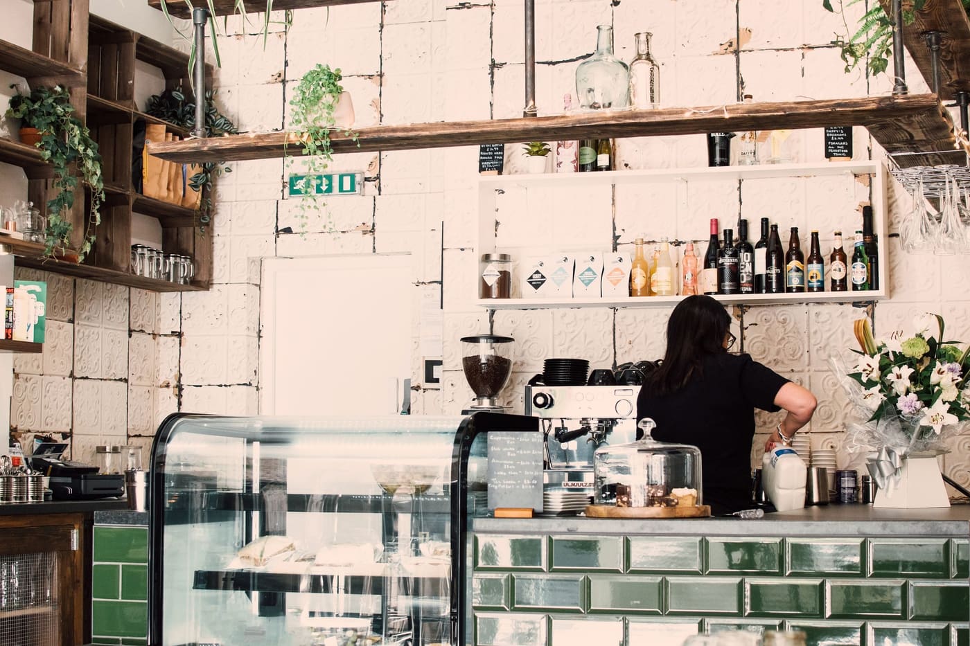 A barista working behind a café counter with green tiles and a cake cabinet
