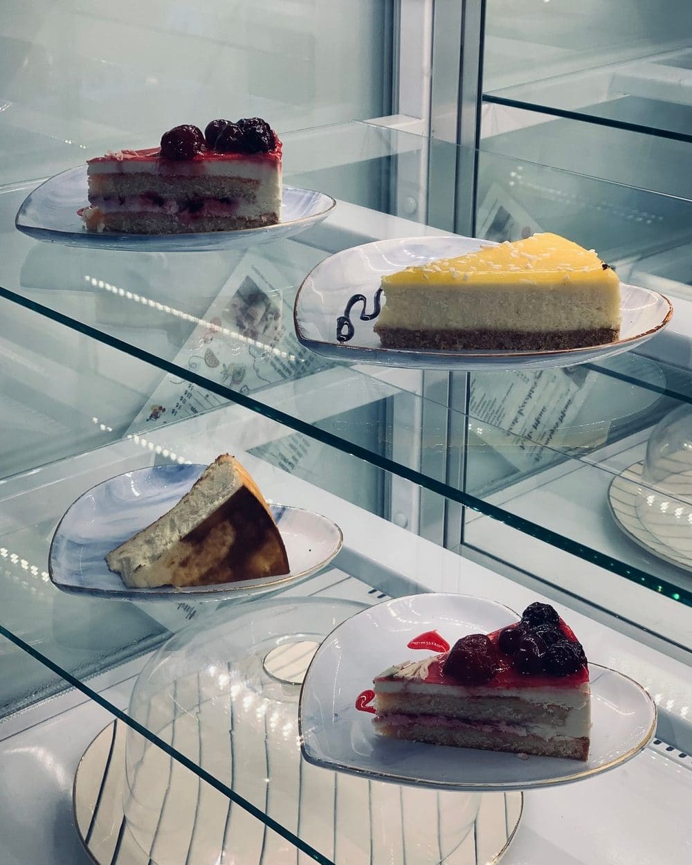Slices of cheesecake and berry cake on glass shelves in the cabinet