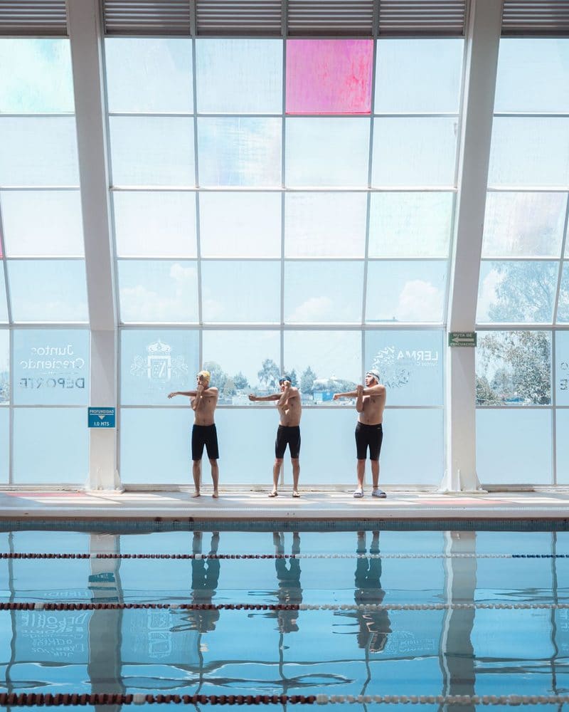 Three swimmers stretching on the edge of an indoor pool in front of tall windows
