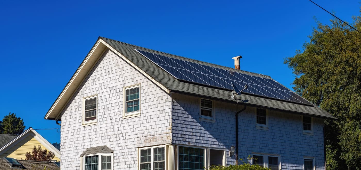 A two-storey weatherboard house with solar panels across the roof under a blue sky