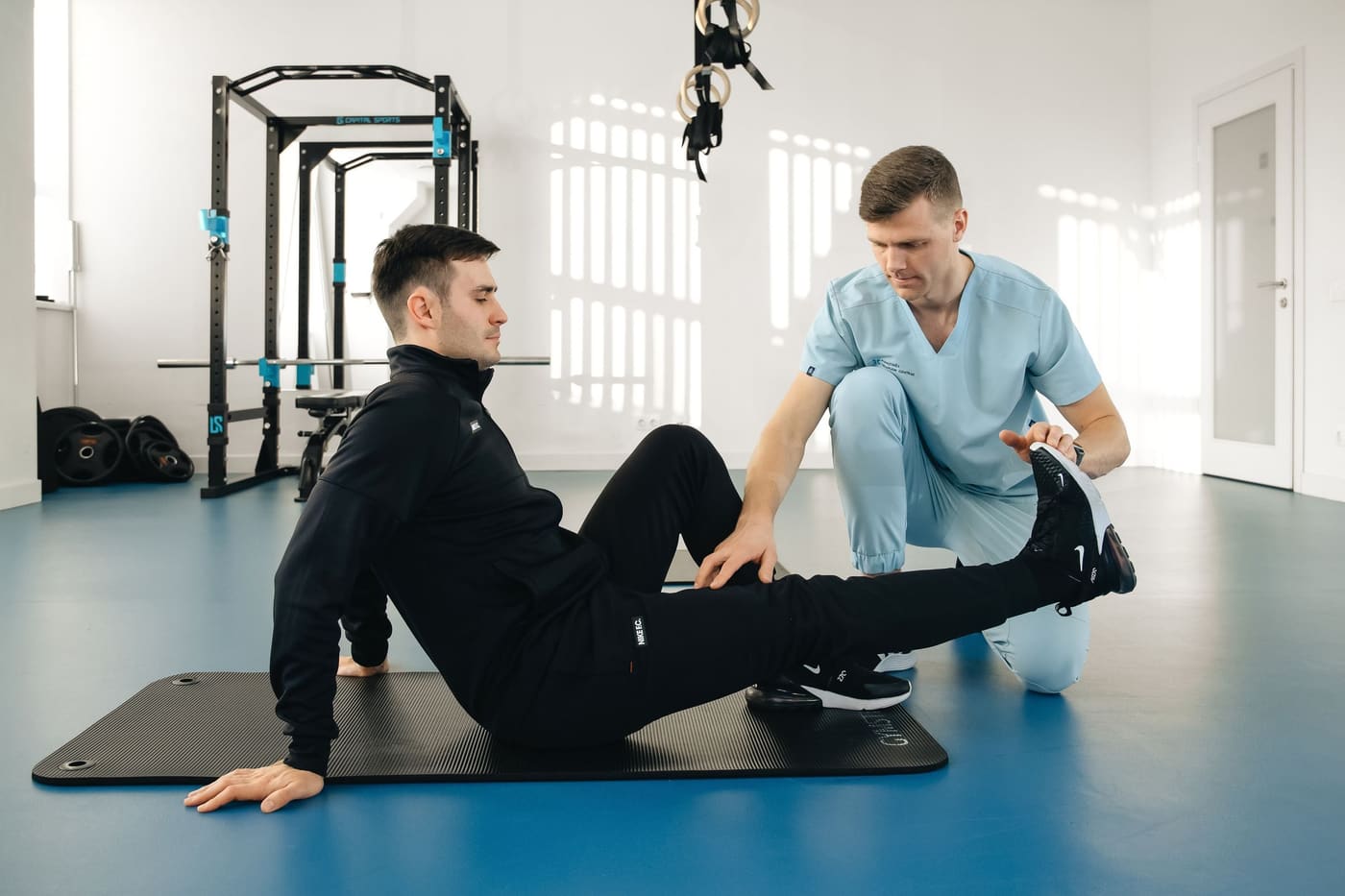 A physiotherapist kneeling beside a man stretching his leg on a mat in a bright gym