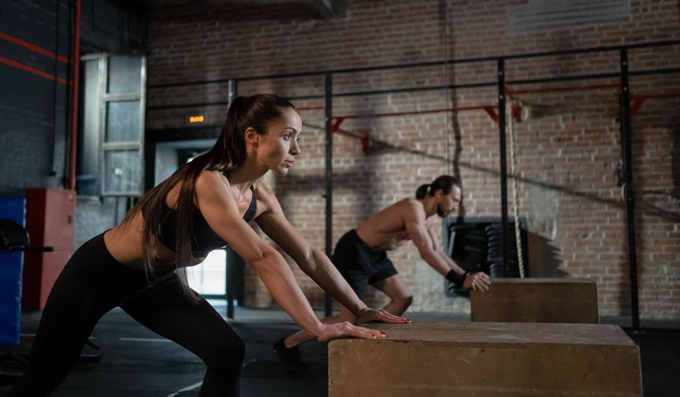 A woman and a man pushing off wooden boxes during a class in a brick-walled gym