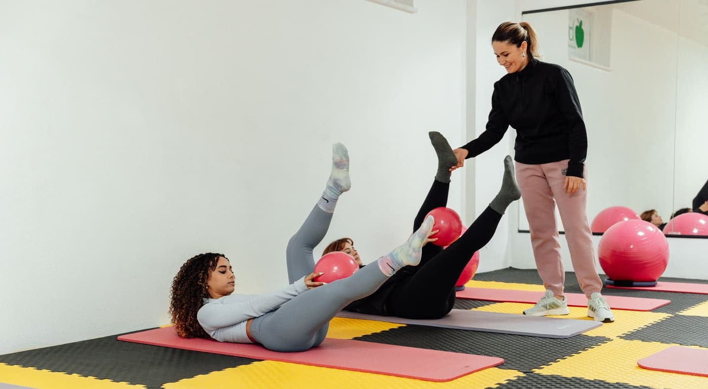 A coach helping two women through a core exercise on mats in a bright studio
