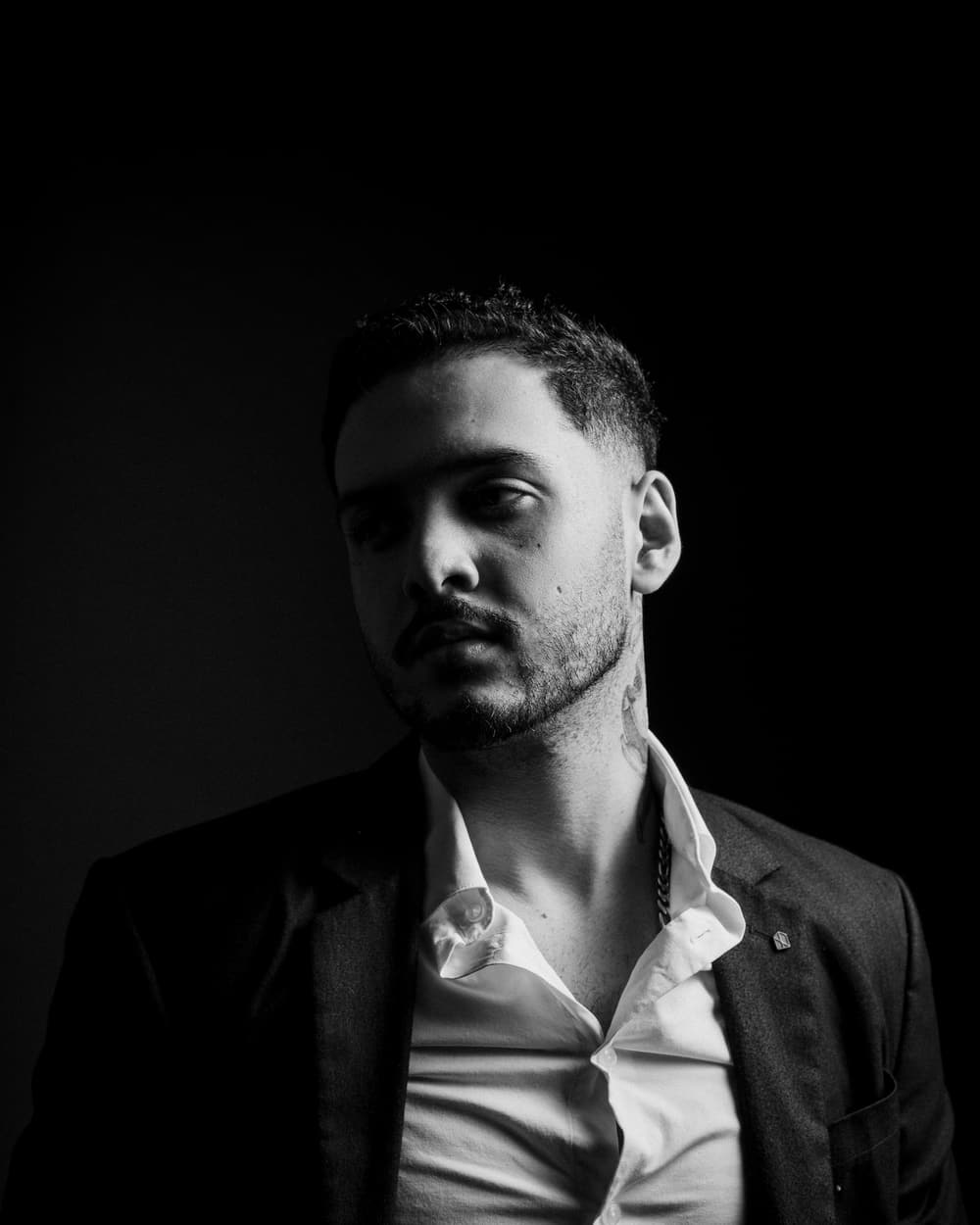 Drew Sample in a dark jacket and open shirt, in black and white against a dark background