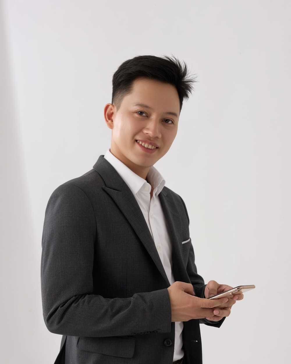 Kai Sample in a dark suit holding a phone, against a white wall