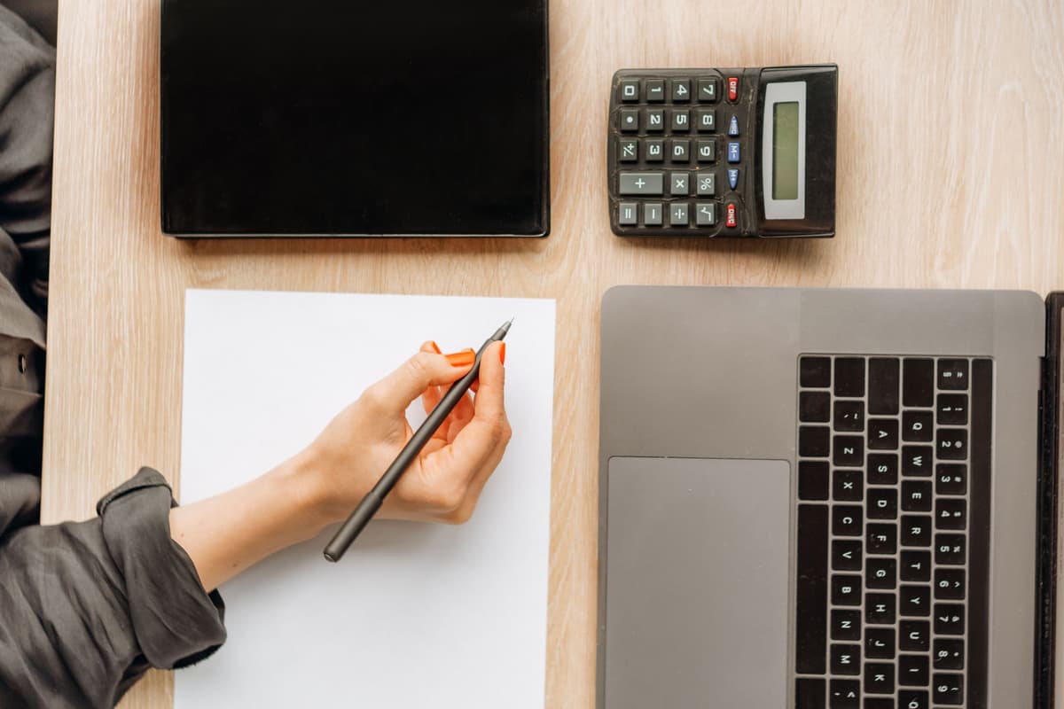 A hand writing on a notepad beside a laptop and a calculator