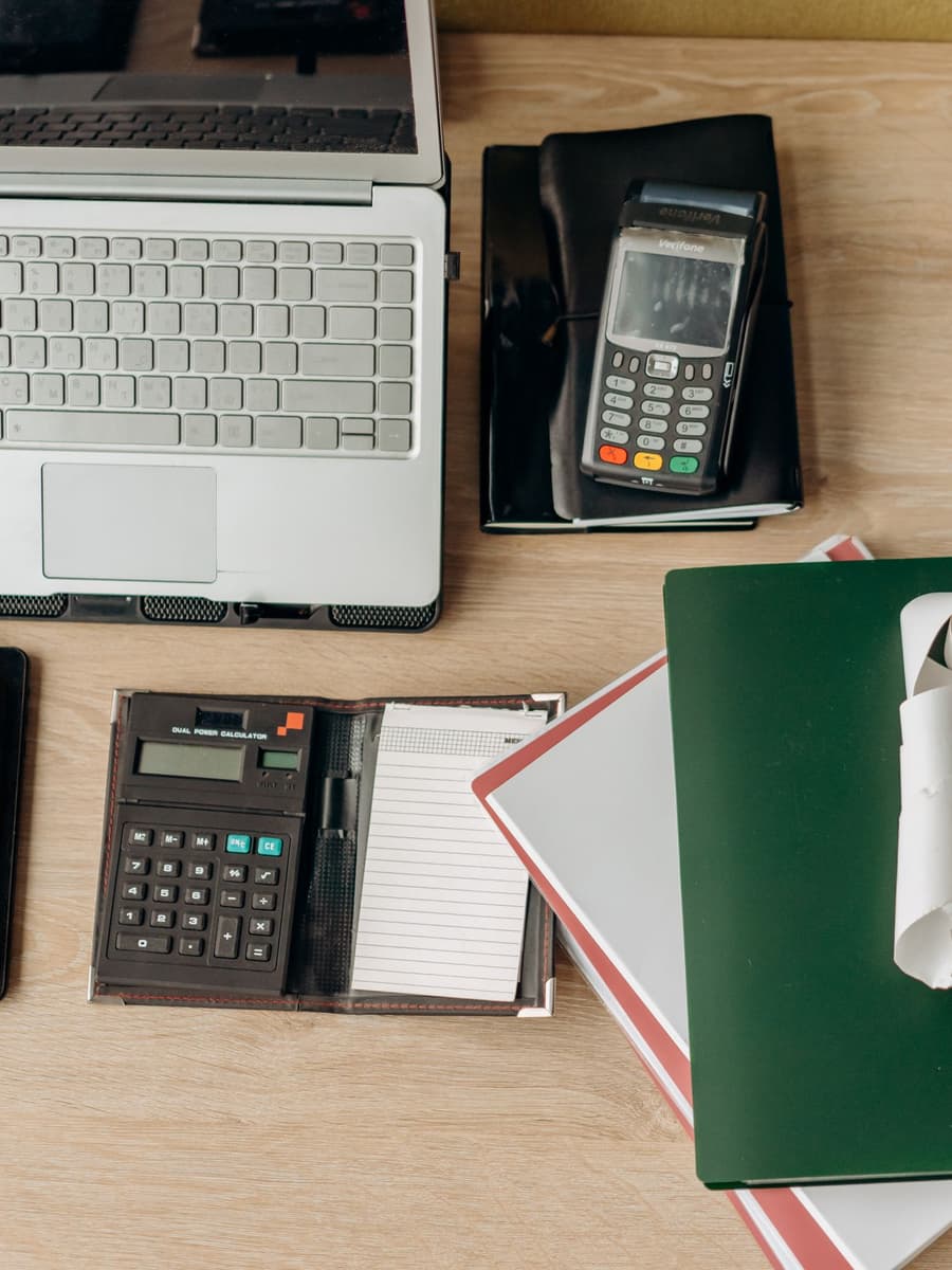 A desk seen from above with a laptop, a card reader, a calculator and folders